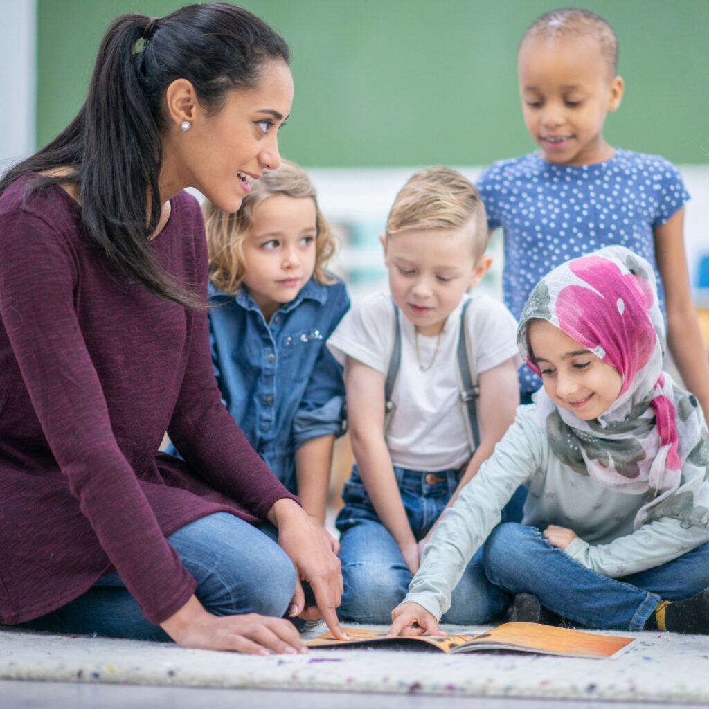 Image shows a school teacher pointing to a book with a group of young children