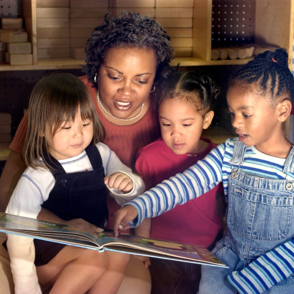 Image shows a teacher looking at a book with three students. One girl is pointing to the book.