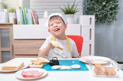 Cute Asian boy wearing chef hat and apron having fun preparing sandwiches, Little kid holding bread while making sandwiches at home, Fun indoor activities for kindergarten kid concept